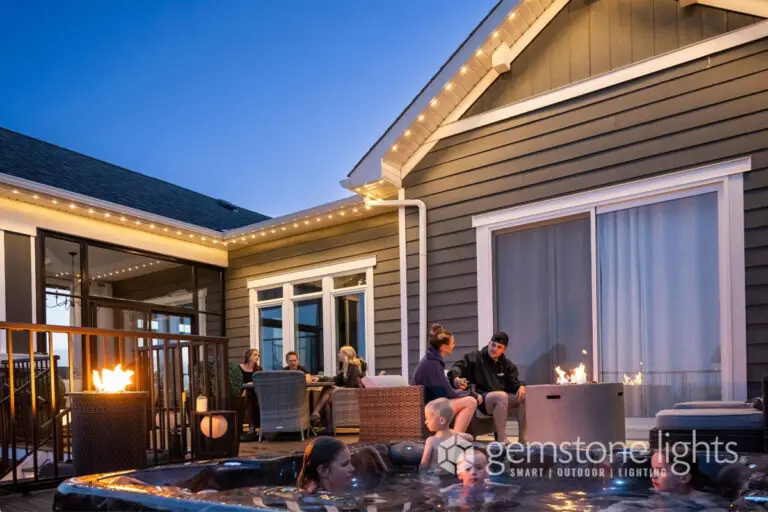 Warm white LED soffit lighting on a home exterior with people gathered around a hot tub and fire pit