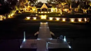 Evening view of a waterfront property with pathway and dock lights leading up to a well-lit backyard and firepit area.