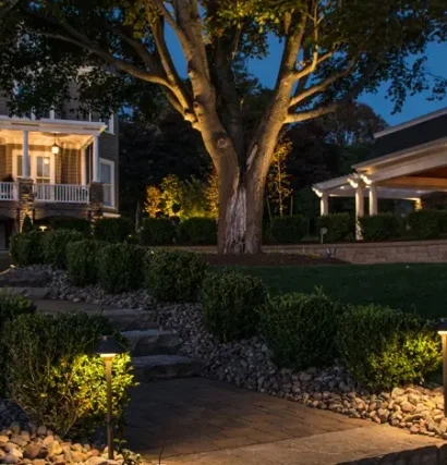 Evening view of a stone pathway and garden lighting leading to a large home with tree uplighting