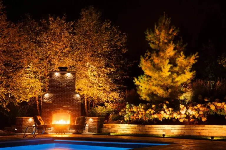 Backyard fireplace area surrounded by trees with warm uplighting, creating an inviting and luxurious nighttime atmosphere.