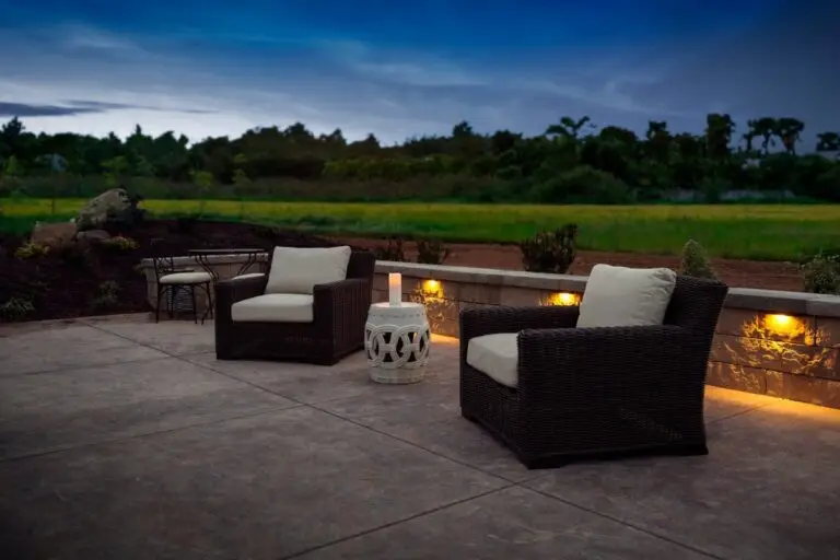 Warm LED hardscape lights installed in a low stone wall create a cozy outdoor seating area with wicker chairs
