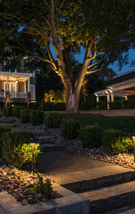 Evening view of a stone pathway and garden lighting leading to a large home with tree uplighting