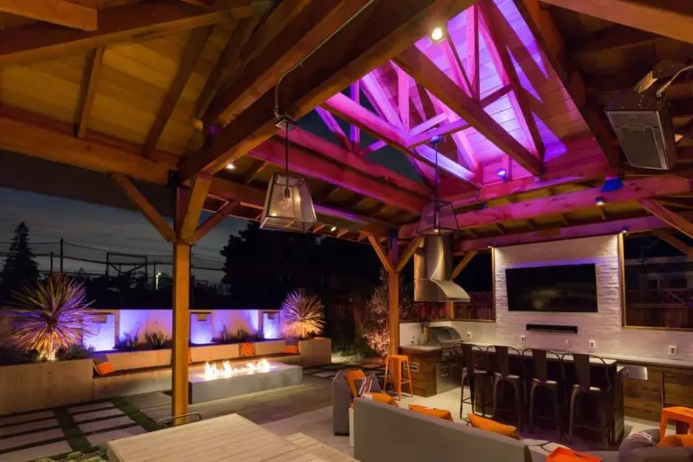 Outdoor kitchen and seating area lit with accent lighting and purple uplights on plants and beams