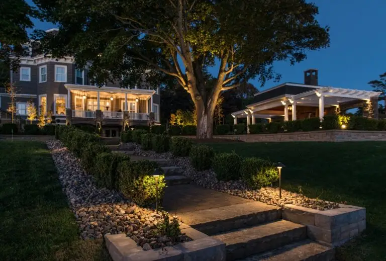 Evening view of a stone pathway and garden lighting leading to a large home with tree uplighting