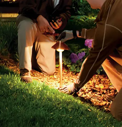 Technicians repairing a landscape lighting fixture in a garden bed at dusk in the Florida Panhandle