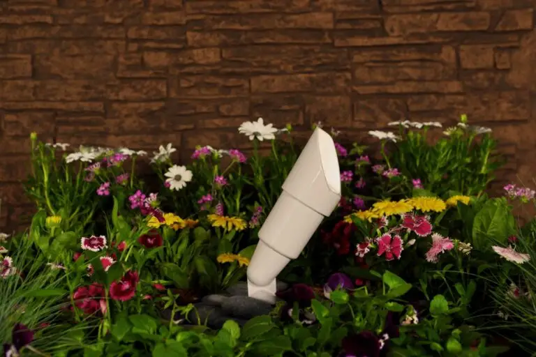 Close-up of a white landscape uplight fixture installed among blooming flowers with a stone wall background.